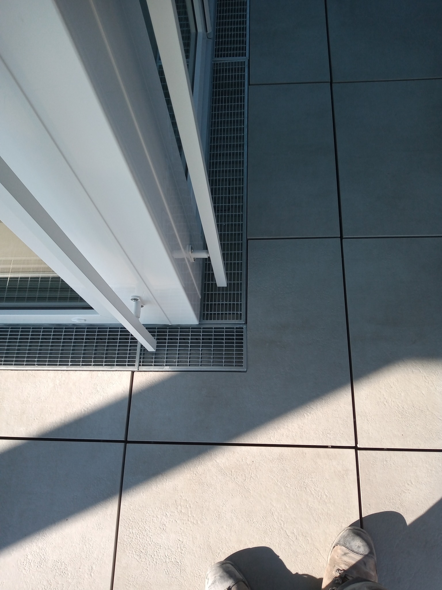 Białe drzwi balkonowe z kratką odpływową w posadzce z jasnych płytek, cień i fragment buta w dolnej części kadru.