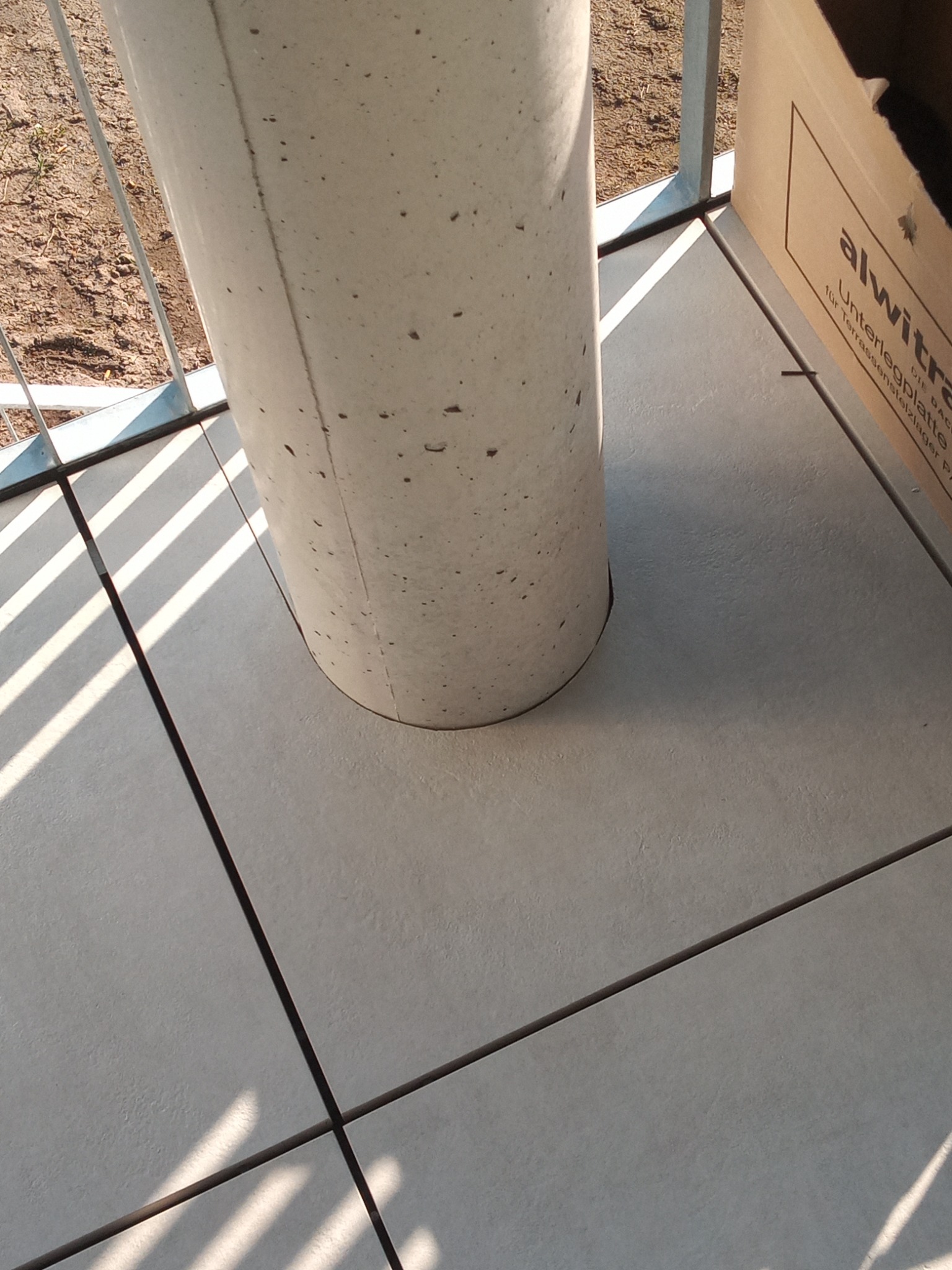 Szare płytki podłogowe na balkonie z betonowym słupem i kartonem w rogu, widok z góry z promieniami słońca.