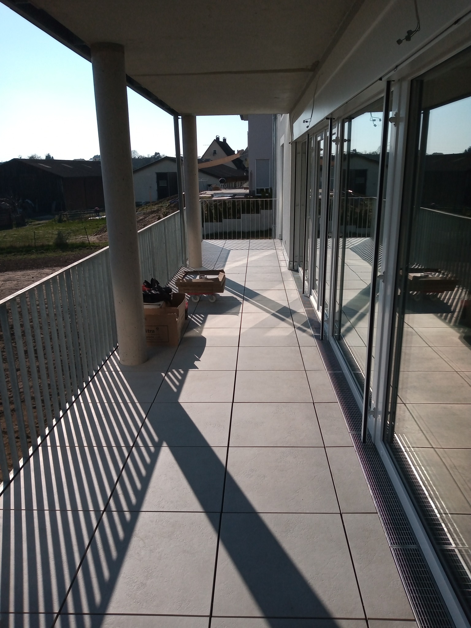 Nowoczesny balkon z szarymi płytkami podłogowymi, metalową balustradą rzucającą cienie i przeszklonymi drzwiami balkonowymi, widoczne kartony i narzędzia.