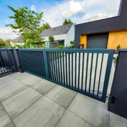 Nowoczesna, grafitowa brama wjazdowa z pionowymi szczeblami, otwierana na posesję z kostki brukowej, z widocznym fragmentem domu w tle.