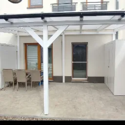 Zadaszony taras z poliwęglanowym dachem, białymi ścianami z desek, pomieszczeniem gospodarczym i zestawem mebli ogrodowych na jasnej, kwadratowej nawierzchni.