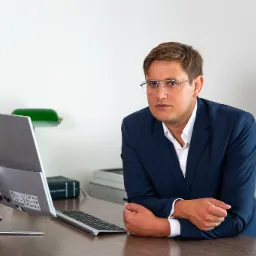 Mężczyzna w okularach i granatowym garniturze opiera się o biurko z komputerem Lenovo, patrząc wprost w obiektyw.