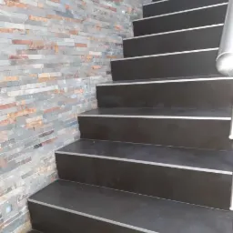 Schody wyłożone ciemnymi płytkami z metalowymi listwami krawędziowymi, ściana obłożona dekoracyjnym kamieniem w odcieniach szarości i brązu, oświetlenie punktowe przy schodach.