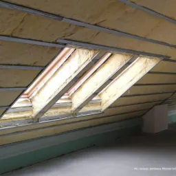 Poddasze z widoczną izolacją z wełny mineralnej, zamontowaną między krokwiami i profilami metalowymi, widoczne okno dachowe w trakcie montażu izolacji, surowy stan wykończenia wnętrza.