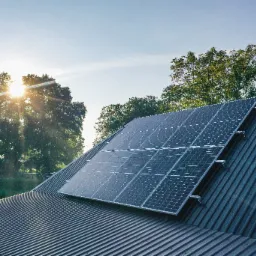 Panele słoneczne na metalowym dachu domu, widok z dołu pod kątem, w tle drzewa i słońce.