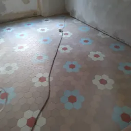 Ułożona podłoga z heksagonalnych płytek w kwiatowy wzór w odcieniach beżu, błękitu i czerwieni w trakcie prac wykończeniowych, widoczne ślady po tynku na ścianach.