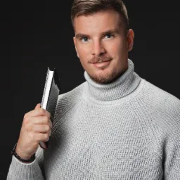 Portret mężczyzny w szarym golfie trzymającego czarny notatnik na ciemnym tle, z subtelnym uśmiechem i bransoletką na nadgarstku.