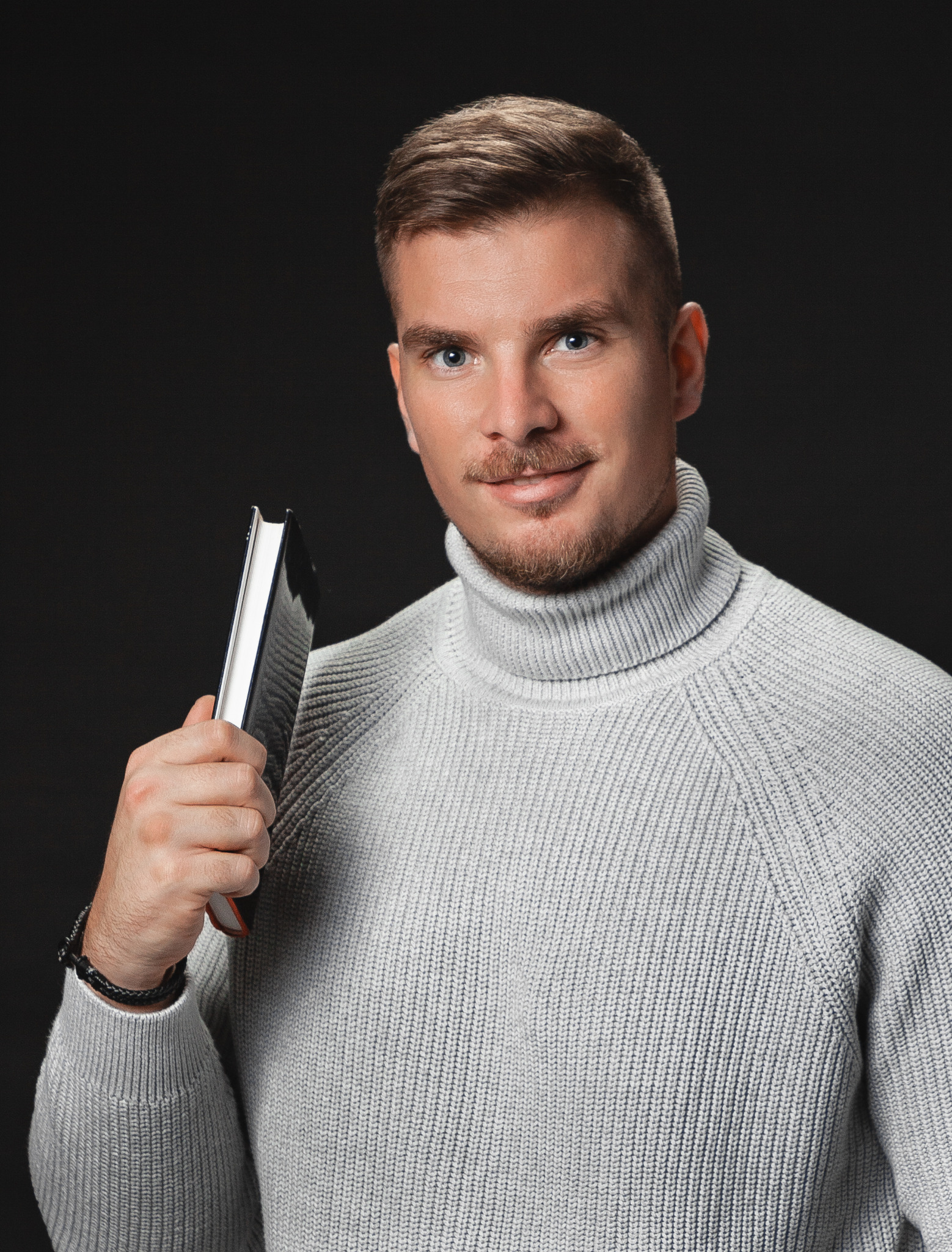 Portret mężczyzny w szarym golfie trzymającego czarny notatnik na ciemnym tle, z subtelnym uśmiechem i bransoletką na nadgarstku.