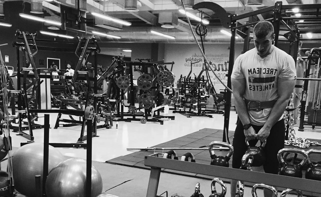 Czarno-białe zdjęcie mężczyzny w pasie kulturystycznym, przygotowującego się do podniesienia kettlebell w dobrze wyposażonej siłowni z różnorodnym sprzętem do ćwiczeń siłowych w tle.