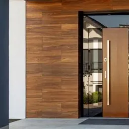 Nowoczesne wejście do domu z brązowymi drzwiami na szklanej ramie, drewnopodobna okładzina elewacyjna i szara kolumna.