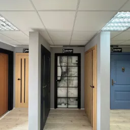 Ekspozycja różnych modeli drzwi wewnętrznych w salonie, widoczne drzwi drewniane, szklane z czarną ramą i niebieskie z kołatką w kształcie lwa.