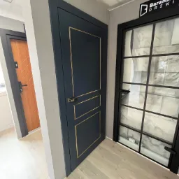 Ekspozycja trzech modeli skrzydeł w salonie: granatowe z złotymi zdobieniami, drewniane z frezowanym wzorem i przeszklone z czarnymi szprosami, podłoga wykończona jasnymi panelami.