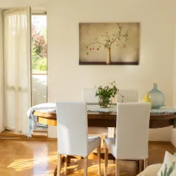 Jasne wnętrze z drewnianym stołem, krzesłami w białych pokrowcach, dekoracjami w postaci wazonów i obrazów, parkiet z połyskującym światłem słonecznym, otwarte drzwi balkonowe z widokiem na zieleń.