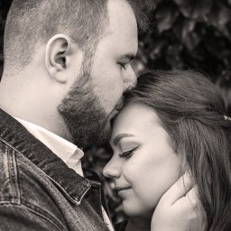 LUX-ON PHOTO Studio mobilne Katarzyna Kęsa - Czarno-białe, intymne zbliżenie: mężczyzna w jeansowej kurtce delikatnie całuje kobietę w czoło, czule obejmując jej twarz dłonią na tle zielonych liści.