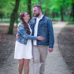 LUX-ON PHOTO Studio mobilne Katarzyna Kęsa - Para zakochanych w Poznaniu spaceruje po parku, ubrana w jeansowe kurtki i białą sukienkę, patrząc na siebie z uśmiechem.