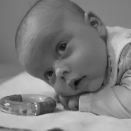 Czarno-białe zdjęcie noworodka leżącego na brzuchu z zabawką, skupionego wzroku skierowanego w górę.