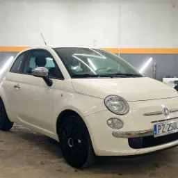 Biały Fiat 500 po kompleksowym myciu, z czarnymi felgami, zaparkowany w dobrze oświetlonym pomieszczeniu myjni, widoczne odbicia światła na lakierze.