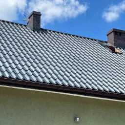 Dom z szarym dachem z dachówki, dwa ceglane kominy, antena telewizyjna i fragment elewacji w kolorze oliwkowym, widoczne brązowe rynny, niebieskie niebo z chmurami w tle.