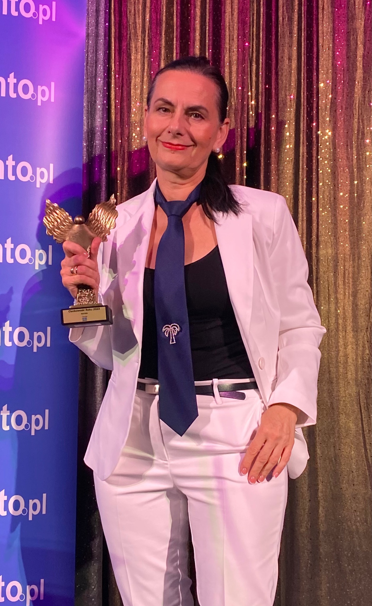 Portret kobiety w białym garniturze, trzymającej statuetkę 'Osobowość Roku 2023' na tle błękitnego baneru i złotej zasłony, z ciemnym krawatem ozdobionym motywem palmy.