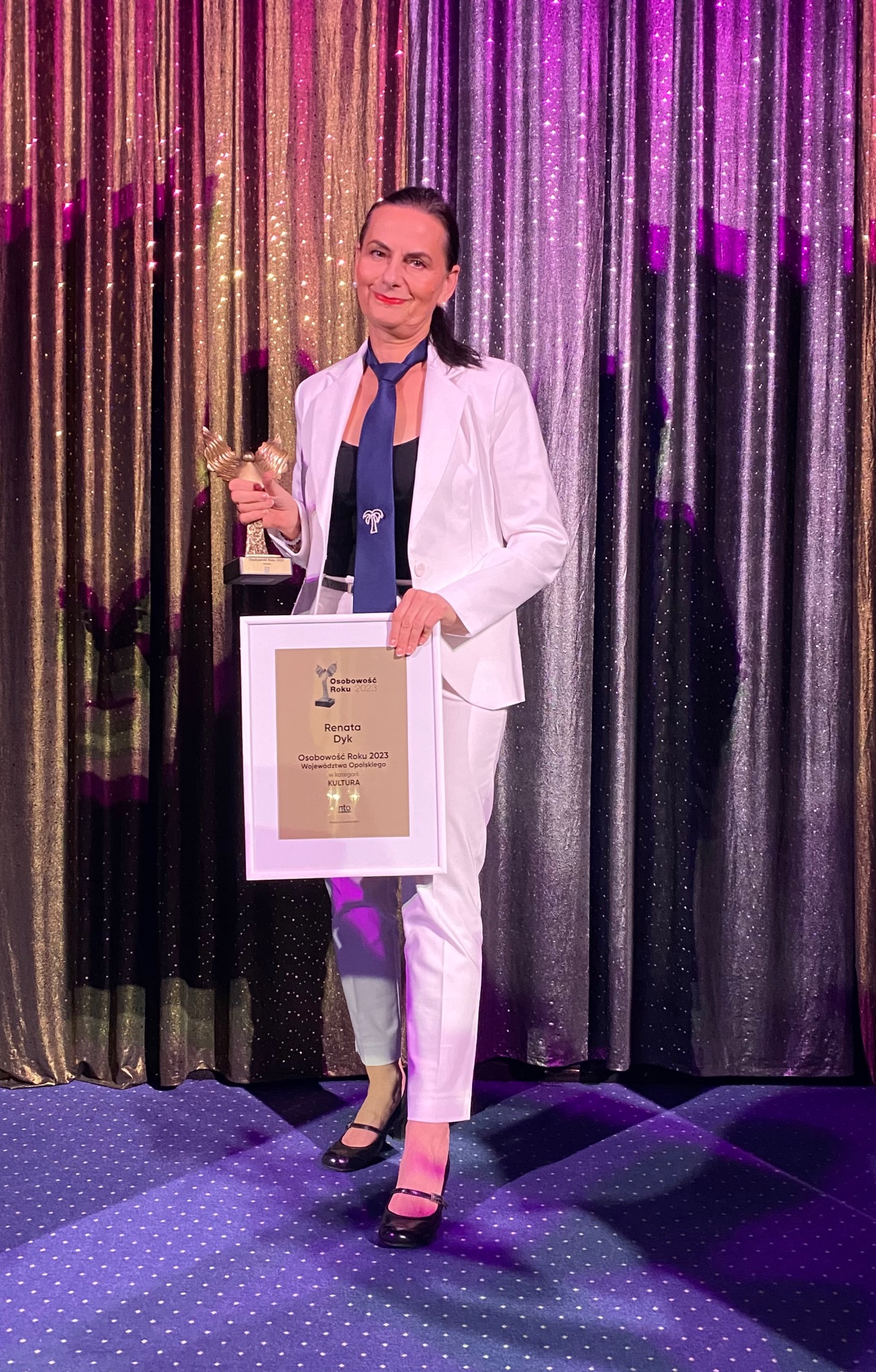 Kobieta w białym garniturze i granatowym krawacie, trzymająca statuetkę i dyplom 'Osobowość Roku 2023' na tle błyszczącej kurtyny.