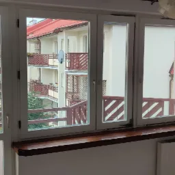 Trzyczęściowe białe okno z drewnianym parapetem, widok na budynki z balkonami i drzewa w jesiennych barwach.