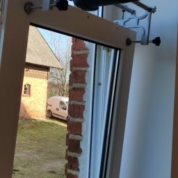 Oknomaniak Marek Pałucki - Frezarka Makita przymocowana do ramy okiennej podczas prac na tle ceglanego budynku i białego samochodu dostawczego.