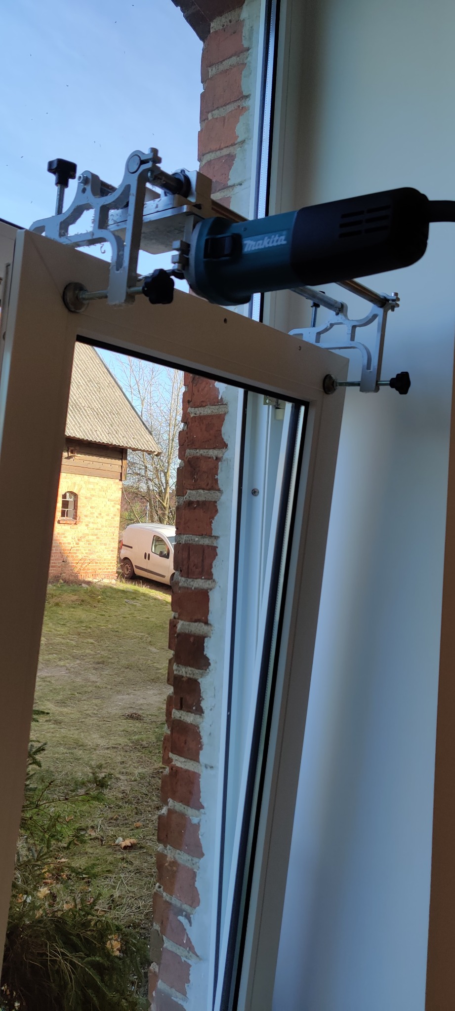 Frezarka Makita przymocowana do ramy okiennej podczas prac na tle ceglanego budynku i białego samochodu dostawczego.