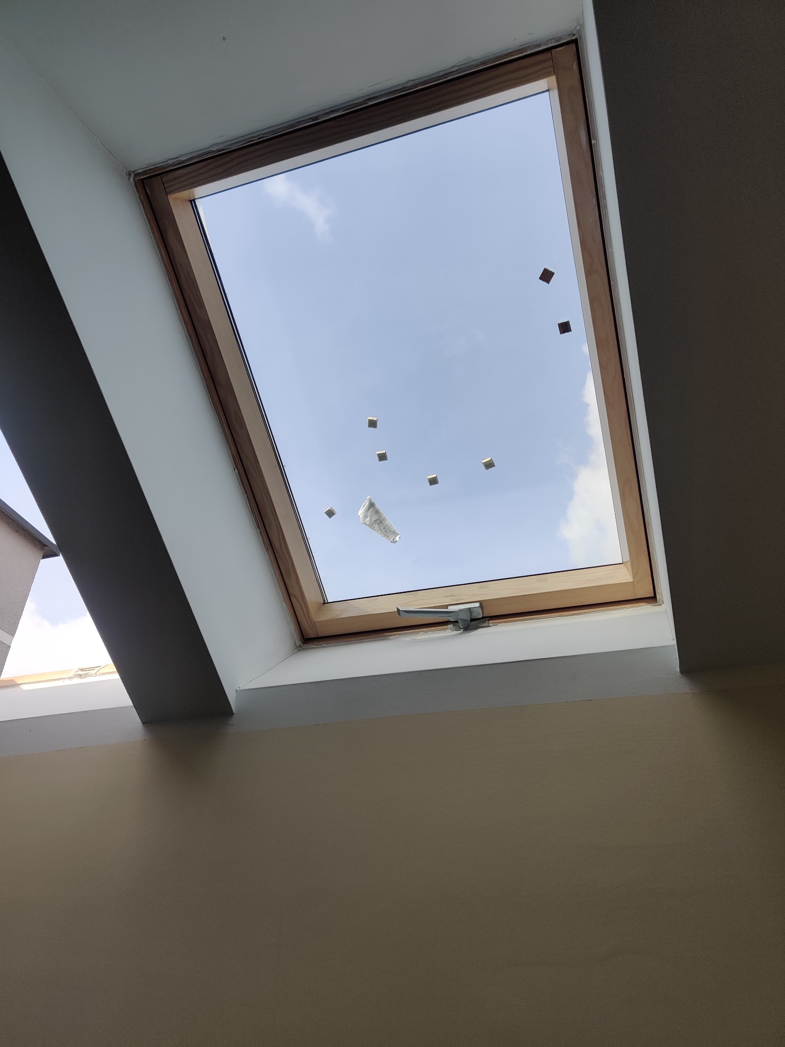 Okno dachowe z drewnianą ramą, widok na błękitne niebo z chmurami, zabezpieczone taśmą malarską po obwodzie szyby, uchwyt okienny widoczny