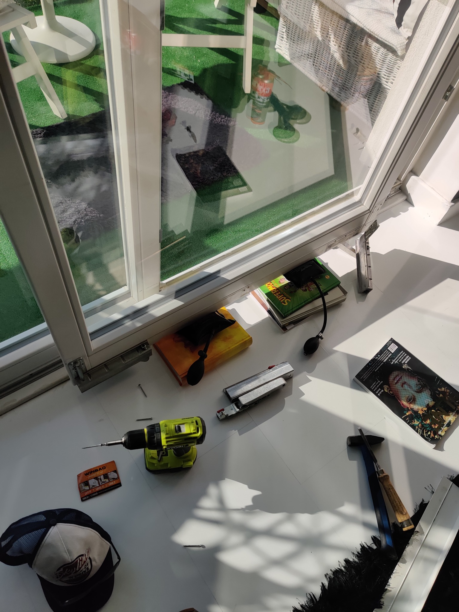 Widok z góry na białe okno z widocznymi narzędziami: wiertarka, młotek, dłuto i poduszka powietrzna do poziomowania okien, oraz czapka z daszkiem i magazyn leżące na parapecie.