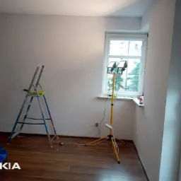 Pokój po odświeżeniu, widoczna drabina, lampa halogenowa na statywie i folia ochronna na podłodze, ściany w neutralnym odcieniu bieli, okno z widokiem na zieleń.