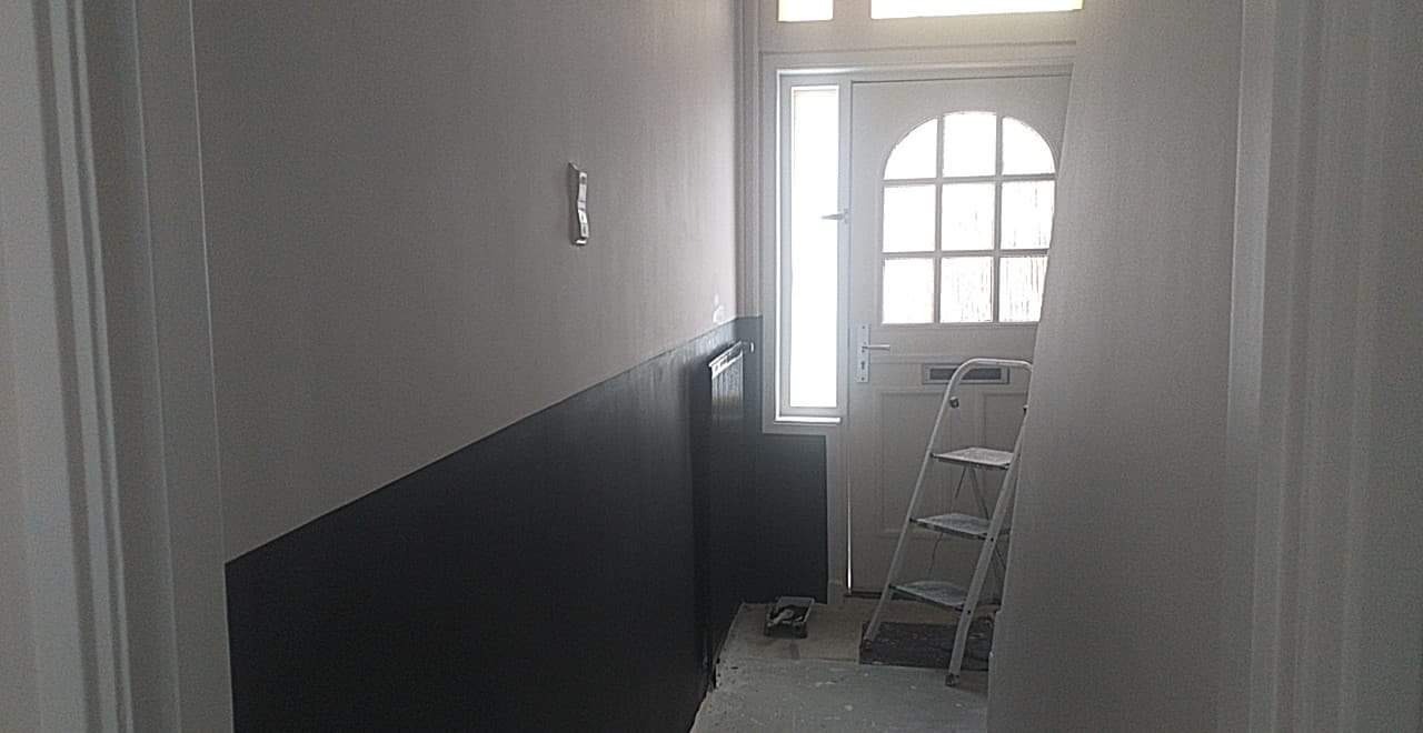 Korytarz w trakcie prac: ściany częściowo pomalowane na szaro i czarno, drabina oparta o drzwi wejściowe, widoczne ślady farby na podłodze.