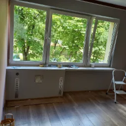 Pokój przed remontem z nowymi oknami, widok na zieleń za oknem, drabinka, panele podłogowe częściowo ułożone, przygotowania do prac wykończeniowych.