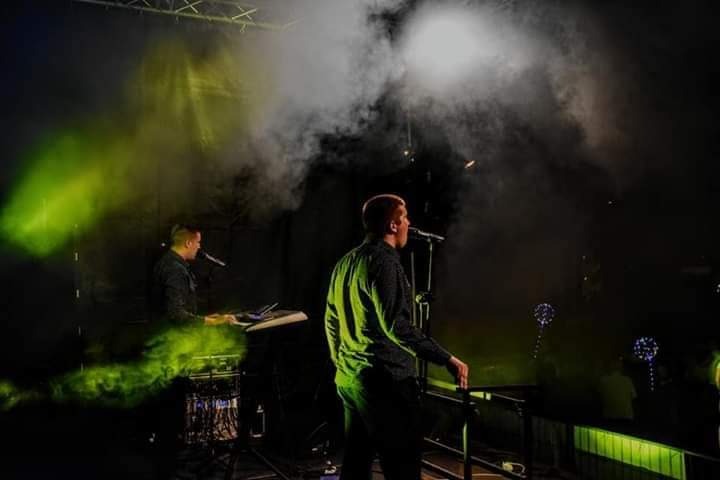 Wokalista z mikrofonem i klawiszowiec w tle, oświetleni zielonym i białym światłem scenicznym, z efektem dymu.