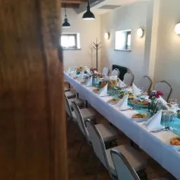 Długi stół nakryty białym obrusem z turkusowym bieżnikiem, przygotowany do uroczystości, z widocznymi potrawami, kwiatami i zastawą, oświetlony wiszącymi lampami w rustykalnym wnętrzu.