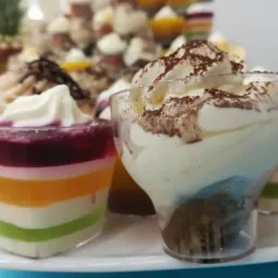 Deser w przezroczystych kubeczkach: tęcza z galaretki i kremu oraz tiramisu posypane kakao, ustawione na białym talerzu na tle innych deserów.