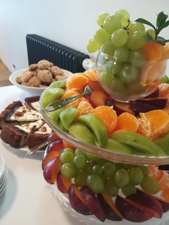 Dwupoziomowa patera z owocami: winogrona, mandarynki, kiwi, śliwki, obok talerz ciasteczek i ciasto na stole.