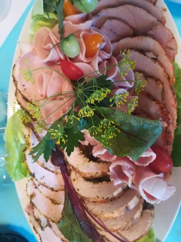 Dekoracyjna kompozycja wędlin, w tym szynki i pieczeni, na białym półmisku, ozdobiona świeżymi ziołami, sałatą i pomidorkami koktajlowymi.