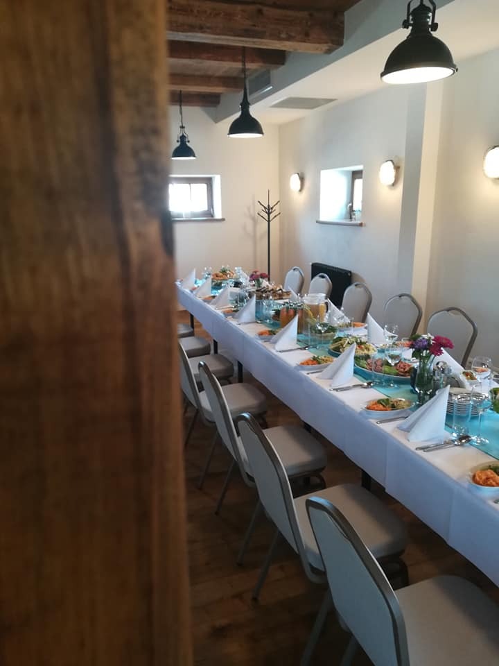 Długi stół nakryty białym obrusem z turkusowym bieżnikiem, przygotowany do uroczystości, z widocznymi potrawami, kwiatami i zastawą, oświetlony wiszącymi lampami w rustykalnym wnętrzu.