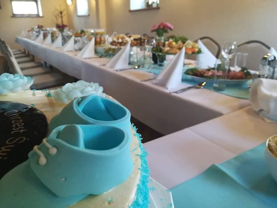 Udekorowany tort z napisem 'Chrzest Święty' i małymi, błękitnymi bucikami z lukru, ustawiony na stole z białym obrusem, nakrytym do uroczystego posiłku. W tle widoczne dania, napoje i dekoracje...