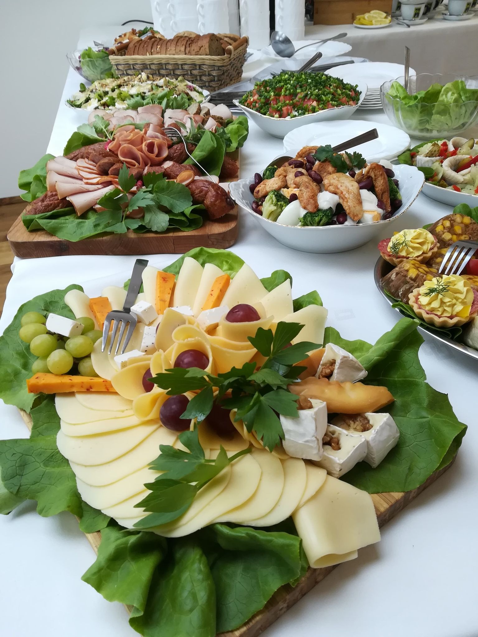 Stół z różnorodnymi potrawami: deski serów z winogronami i zieloną sałatą, sałatki warzywne i mięsne, pieczywo w koszyku, tartaletki z kremem, całość na białym obrusie.