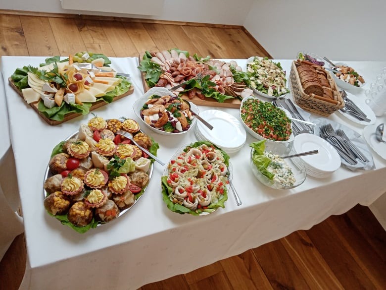 Stół z białym obrusem zastawiony różnorodnymi przekąskami: deski serów i wędlin, sałatki w szklanych misach, tartaletki na srebrnej tacy, pieczywo w koszyku wiklinowym, talerze i sztućce.