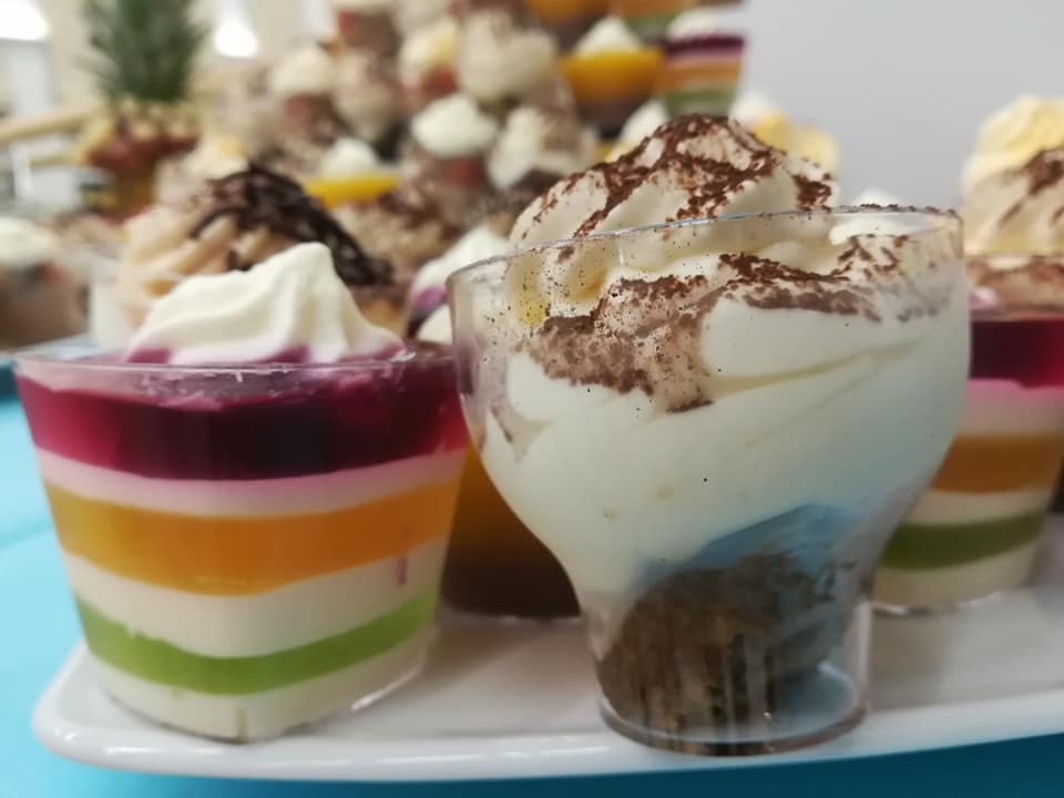 Deser w przezroczystych kubeczkach: tęcza z galaretki i kremu oraz tiramisu posypane kakao, ustawione na białym talerzu na tle innych deserów.