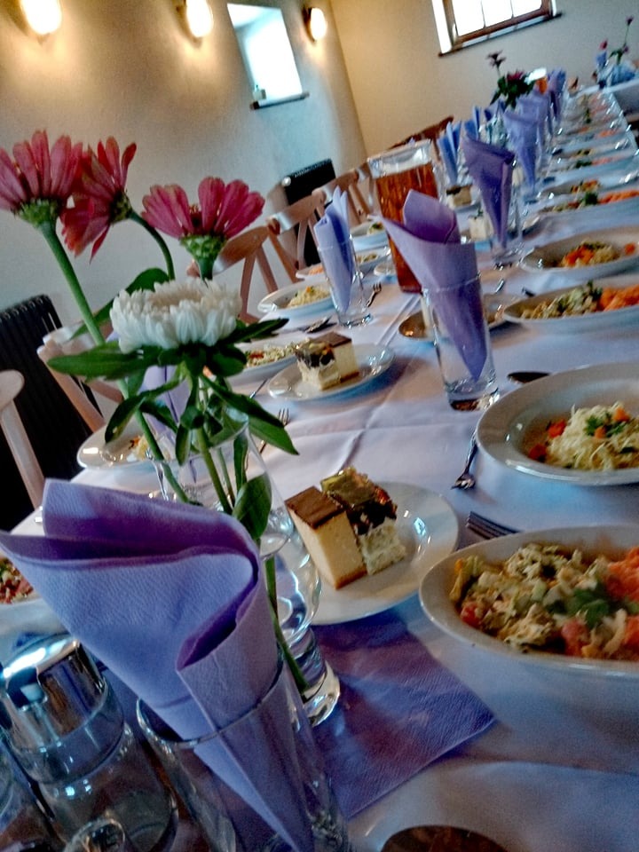 Ustawiony długi stół z białym obrusem, dekoracjami kwiatowymi w wazonach, naczyniami z sałatkami, deserami i fioletowymi serwetkami, przygotowany na przyjęcie.