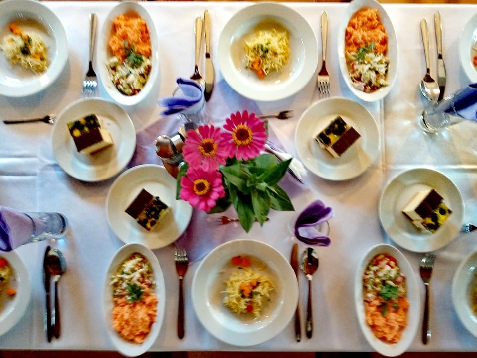 Ustawiony stół z potrawami: sałatki w owalnych miskach, desery czekoladowe z owocami na talerzykach, makaron z warzywami na okrągłych talerzach, sztućce, serwetki i bukiet różowych kwiatów w wazonie.