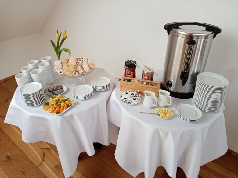 Przygotowany stół w Boguchwale z ciastami, owocami, kawą, herbatą, dzbankami na mleko i podgrzewaczem wody, nakryty białym obrusem, gotowy na przyjęcie gości.
