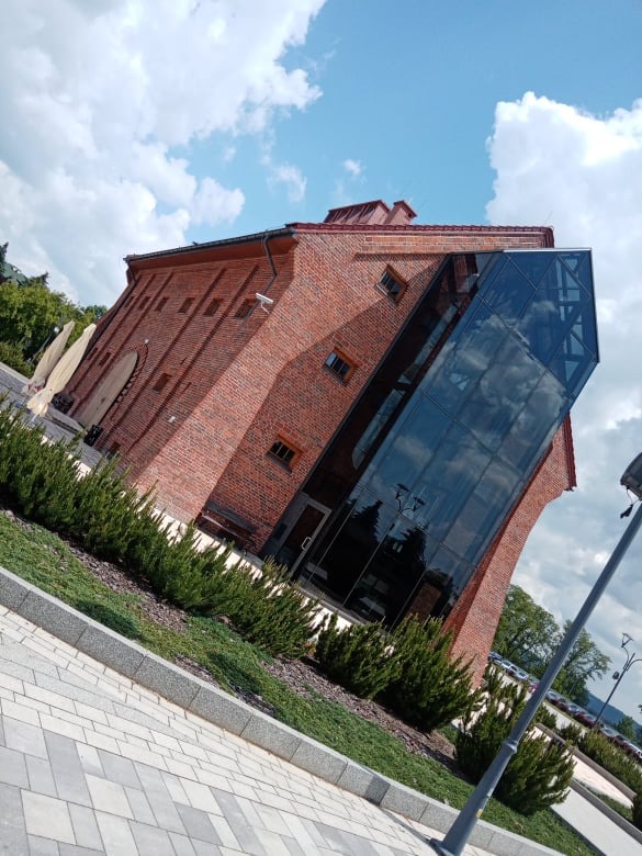 Ceglany budynek z nowoczesną, szklaną dobudówką w kształcie ostrosłupa, otoczony zielenią i brukowanym chodnikiem, widok z dołu w słoneczny dzień.