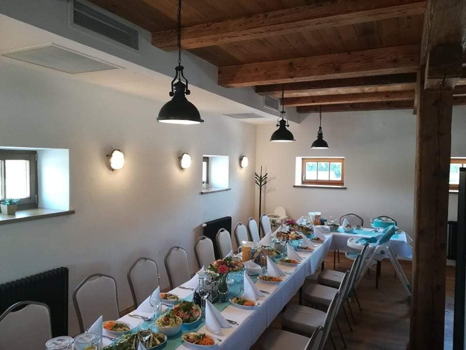 Uroczysty stół z dekoracjami kwiatowymi i zastawą, ustawiony w przestronnej sali z drewnianym stropem i wiszącymi lampami w stylu industrialnym. Widoczne potrawy w misach, szklanki i serwetki.