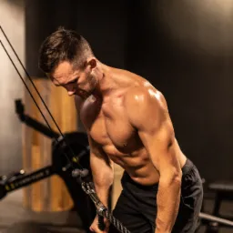 Mężczyzna bez koszulki, z kroplami potu na ciele, ćwiczy na siłowni, trzymając linkę wyciągu, z widocznym tatuażem oka w trójkącie na dłoni.