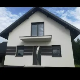 Nowoczesny dom jednorodzinny z białą elewacją, grafitową stolarką okienną i drzwiami z elementami stali nierdzewnej, widoczne oświetlenie zewnętrzne pod dachem.