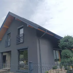 Nowoczesny dom jednorodzinny z szarą elewacją i grafitowymi detalami, widok z boku przez metalowe ogrodzenie, z elementami drewnianymi pod dachem.